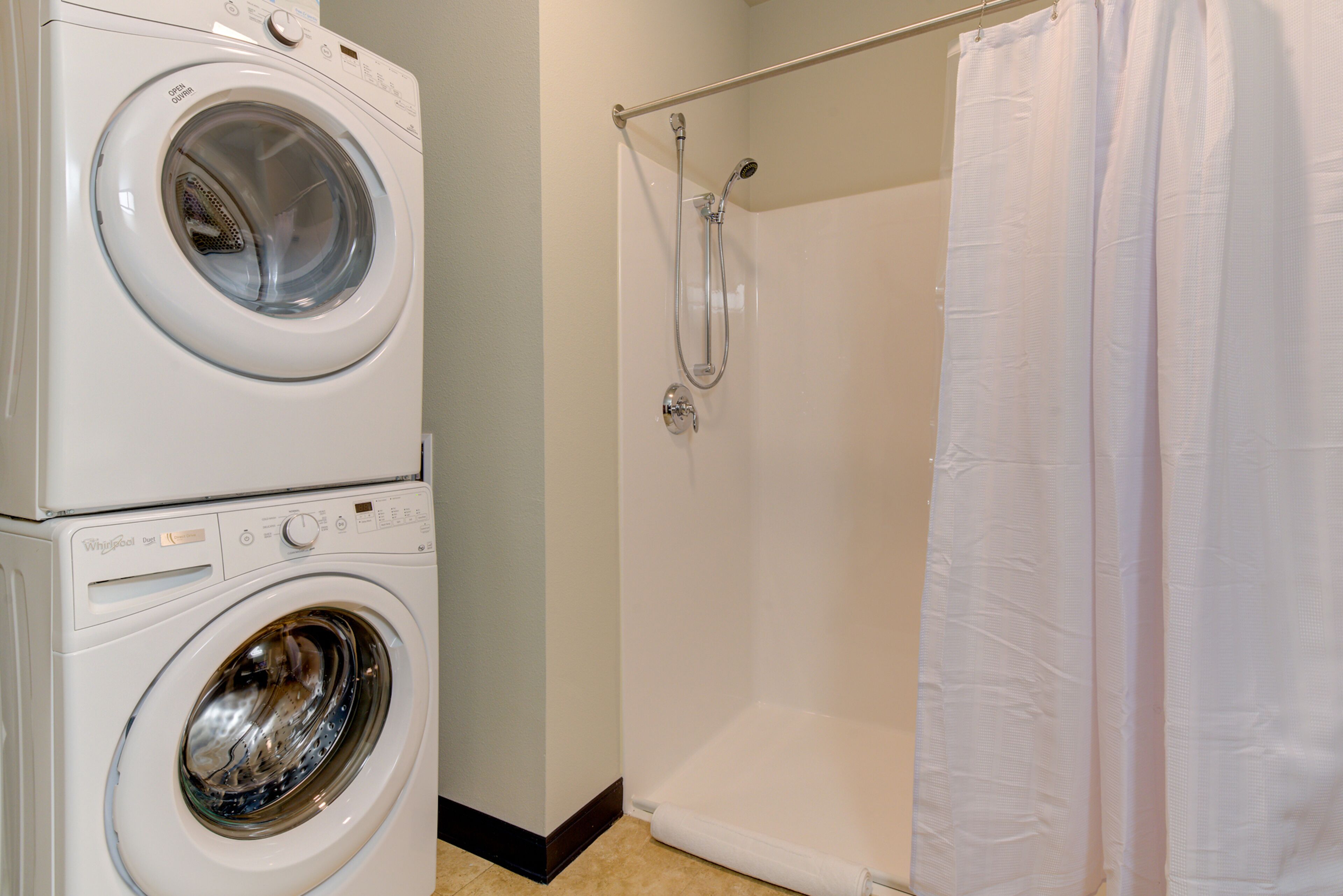 A laundry area with a front-loading washer and dryer, a shower with a glass door, and a white shower curtain.
