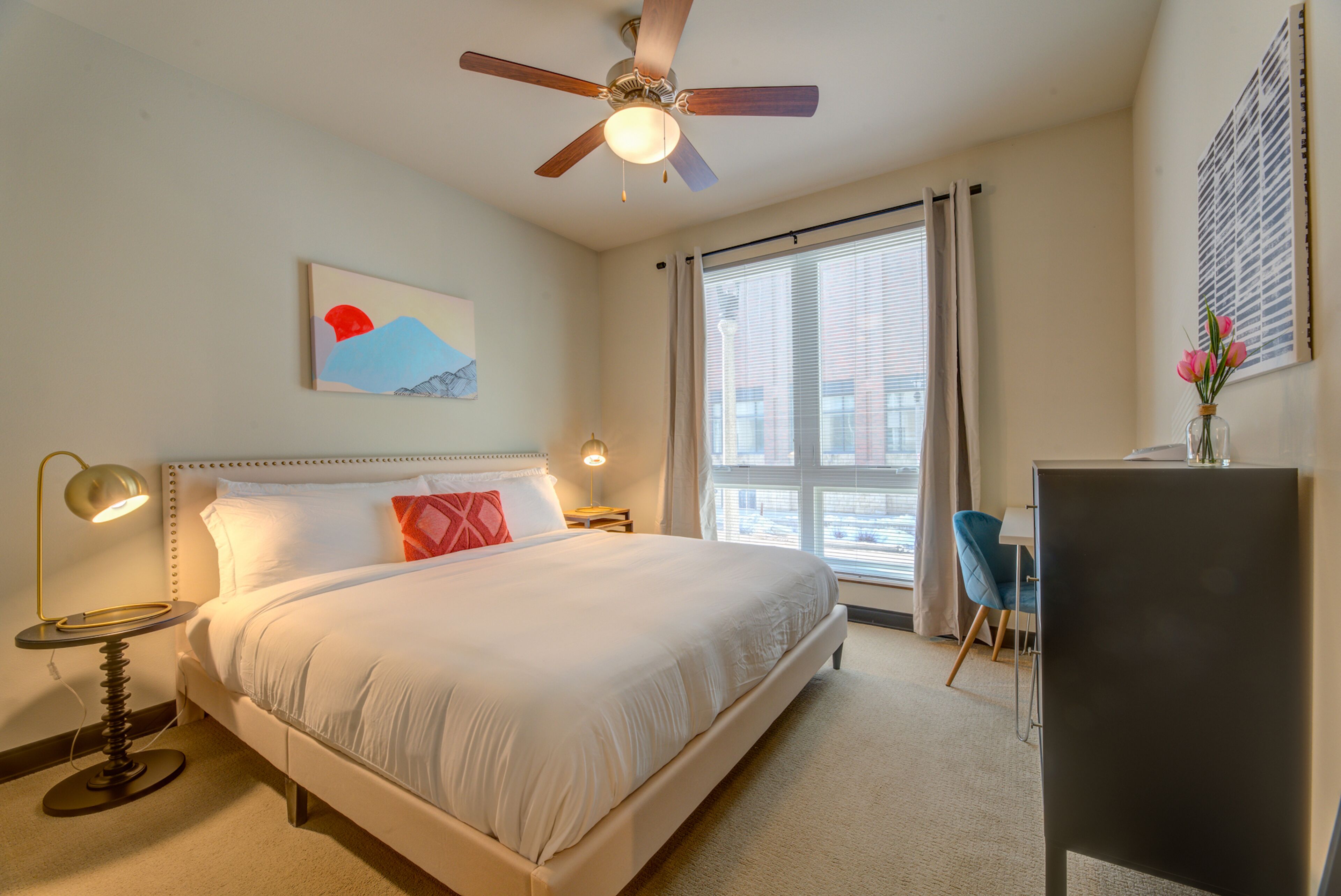 A hotel room with a large bed, a ceiling fan, a desk with a chair, a window with curtains, and a painting on the wall.