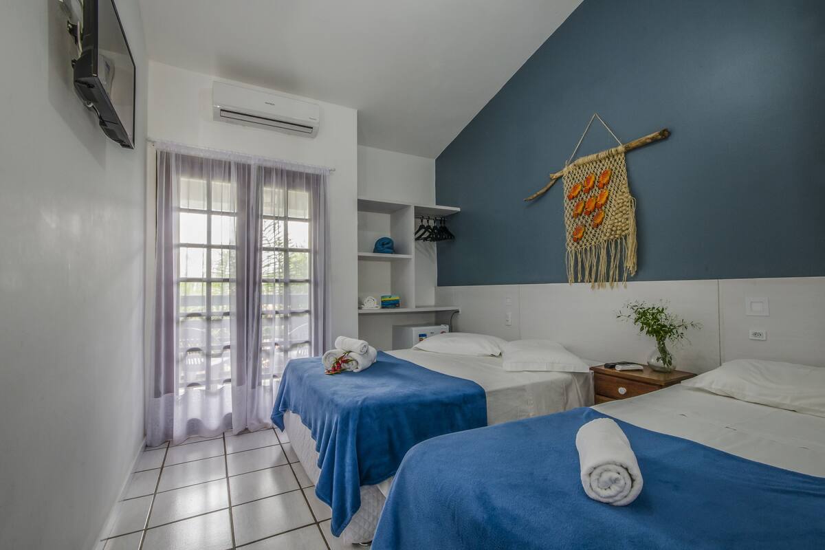 A hotel room with two beds, a small table, a wall-mounted TV, a shelf with personal items, and a window with curtains.