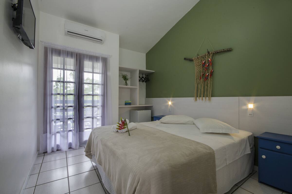 A hotel room with a bed, a blue nightstand, a wall-mounted air conditioner, a shelf with a plant, and a window with sheer curtains.