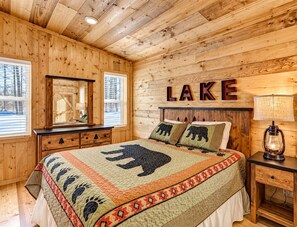 Un dormitorio tipo cabaña con techo de madera, una cama con colcha de estampados de animales, un aparador, una mesita de noche y una lámpara de pared.
