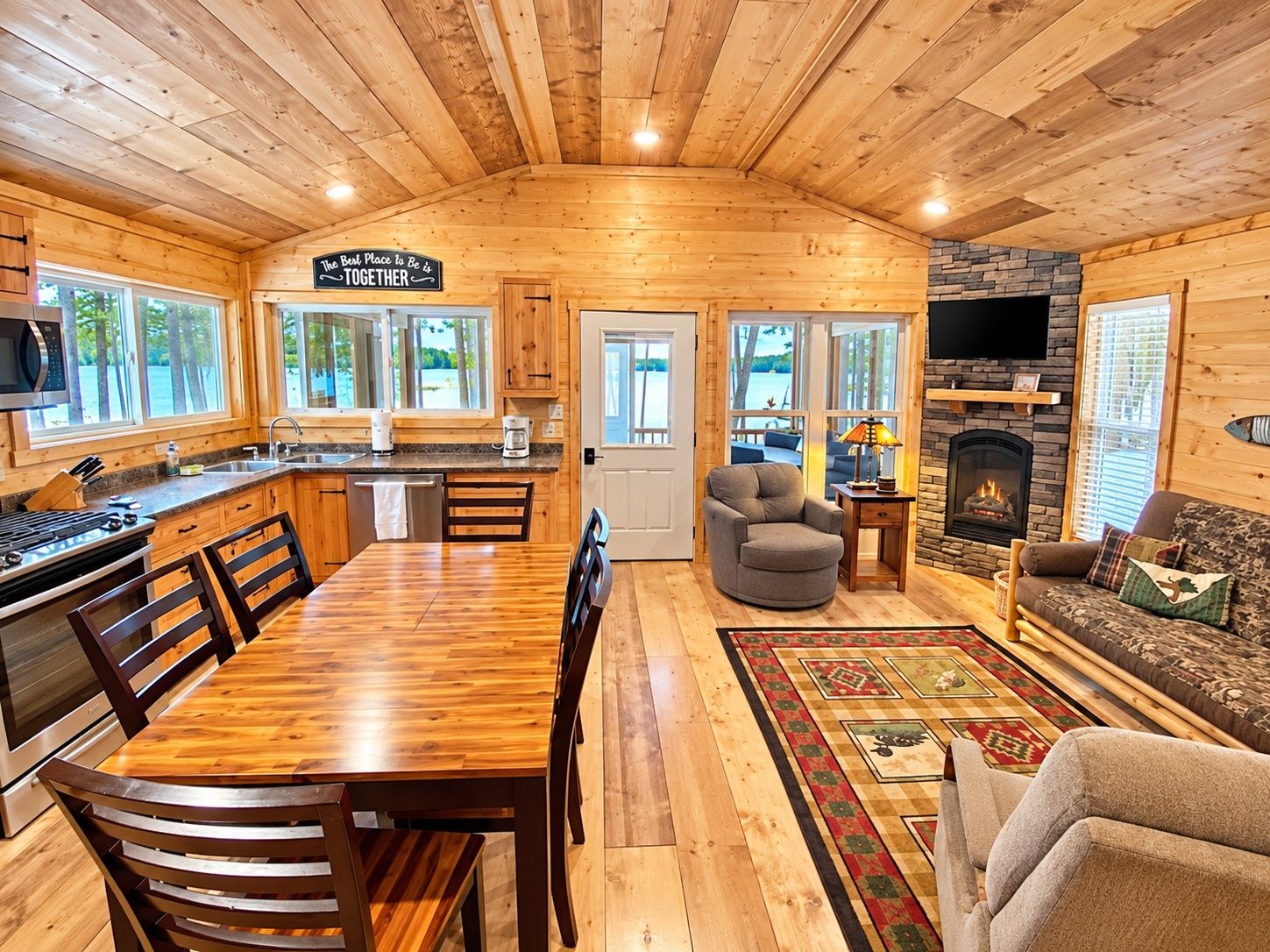 A cabin interior with a kitchen, dining area, and living room, featuring wooden walls and a stone fireplace.