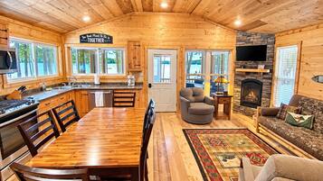 A cabin interior with a kitchen, dining area, and living room, featuring wooden walls and a stone fireplace.