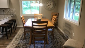 A dining area with a wooden table, four chairs, a chandelier, a clock, and a window with a view of greenery. - Nagawicka Woods-Beautiful Lake Country Ranch Home (Delafield)