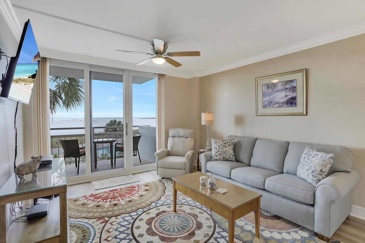 A living room with a sofa, coffee table, and a ceiling fan. There is a TV mounted on the wall and a window with a view of the beach.