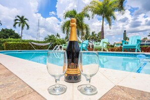 A bottle of Brut Cutte champagne and two glasses on a poolside table with a view of palm trees and a hammock in the background. - Pet-friendly Two-story Home With Pool, Spa, and Grill (Marco Island)