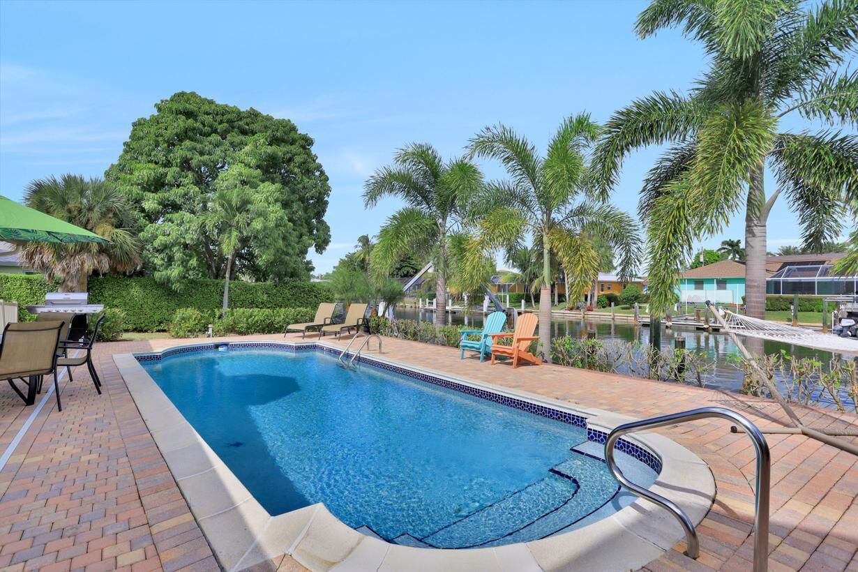 A rectangular swimming pool with a tiled edge, surrounded by a brick patio. There are lounge chairs and a table with a green umbrella.