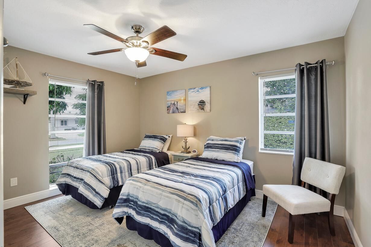 A bedroom with two beds, a ceiling fan, a window with curtains, and a bench.