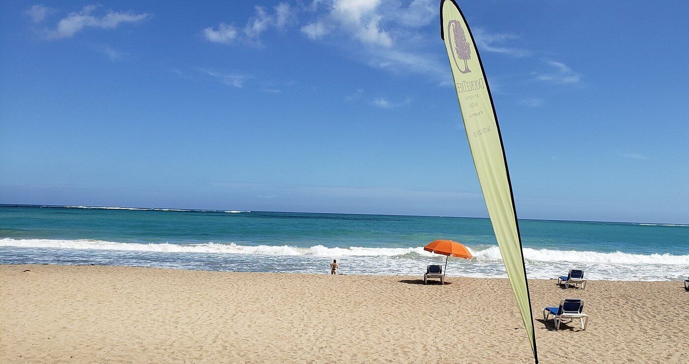Una playa con una bandera, una persona caminando y sillas de playa con una sombrilla.