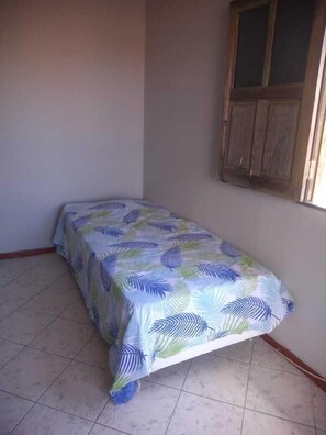 A small, unadorned room with a single bed, plain walls, and a window with wooden shutters.