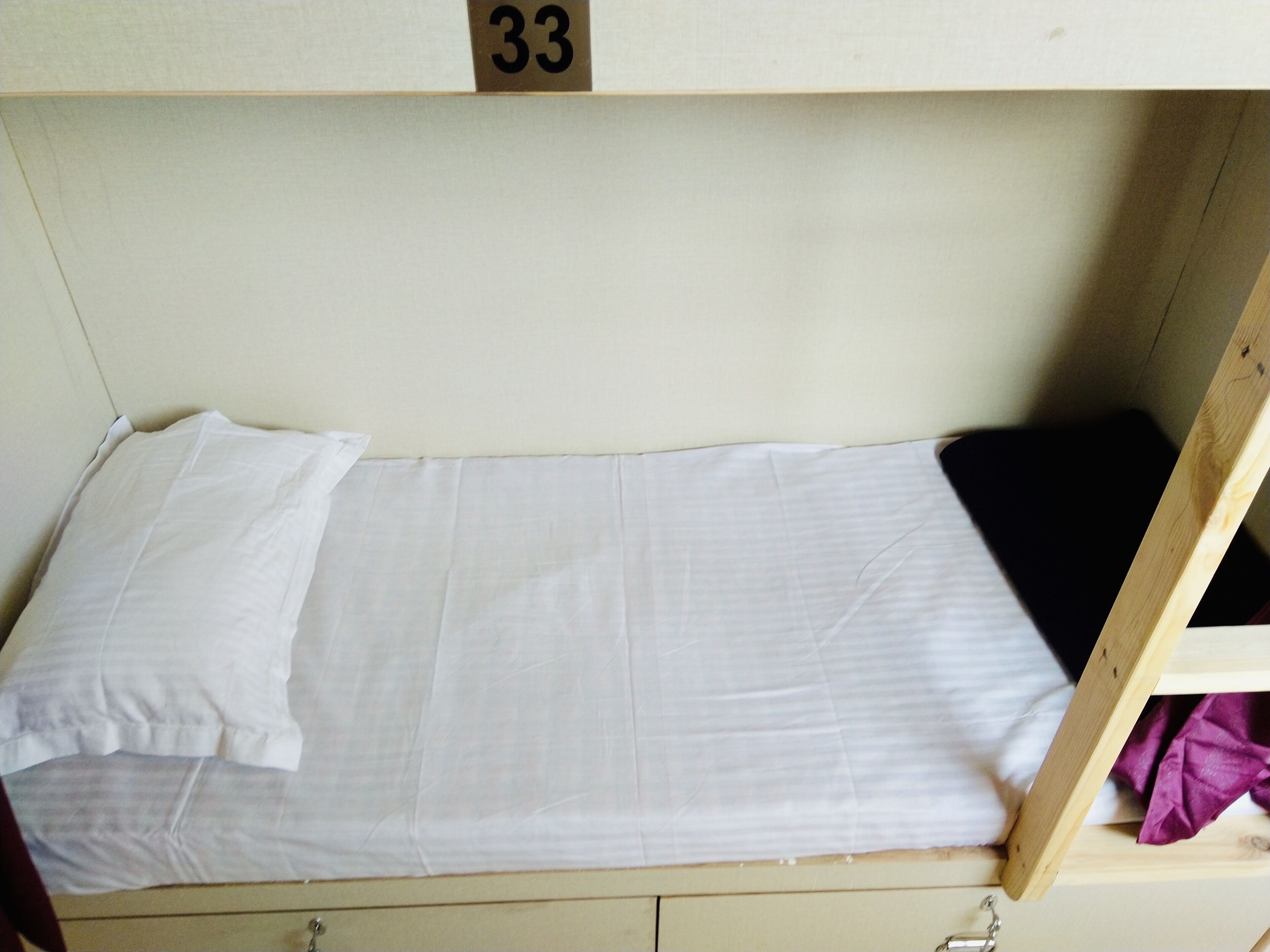 Un lit superposé avec un matelas blanc et un oreiller, une tête de lit en bois portant le numéro 33, et une petite étagère en bois recouverte d’un tissu violet.