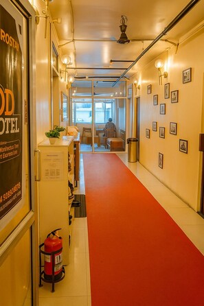 A hotel corridor with a red carpet, a fire extinguisher, framed pictures on the walls, and a person working at a desk in the background.