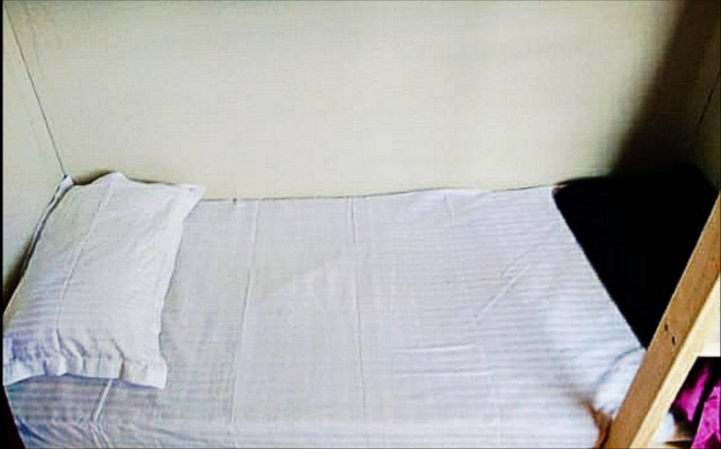 Een eenvoudig bed met een platte matras en witte lakens in een kamer met een effen wand en een houten hoofdbord.