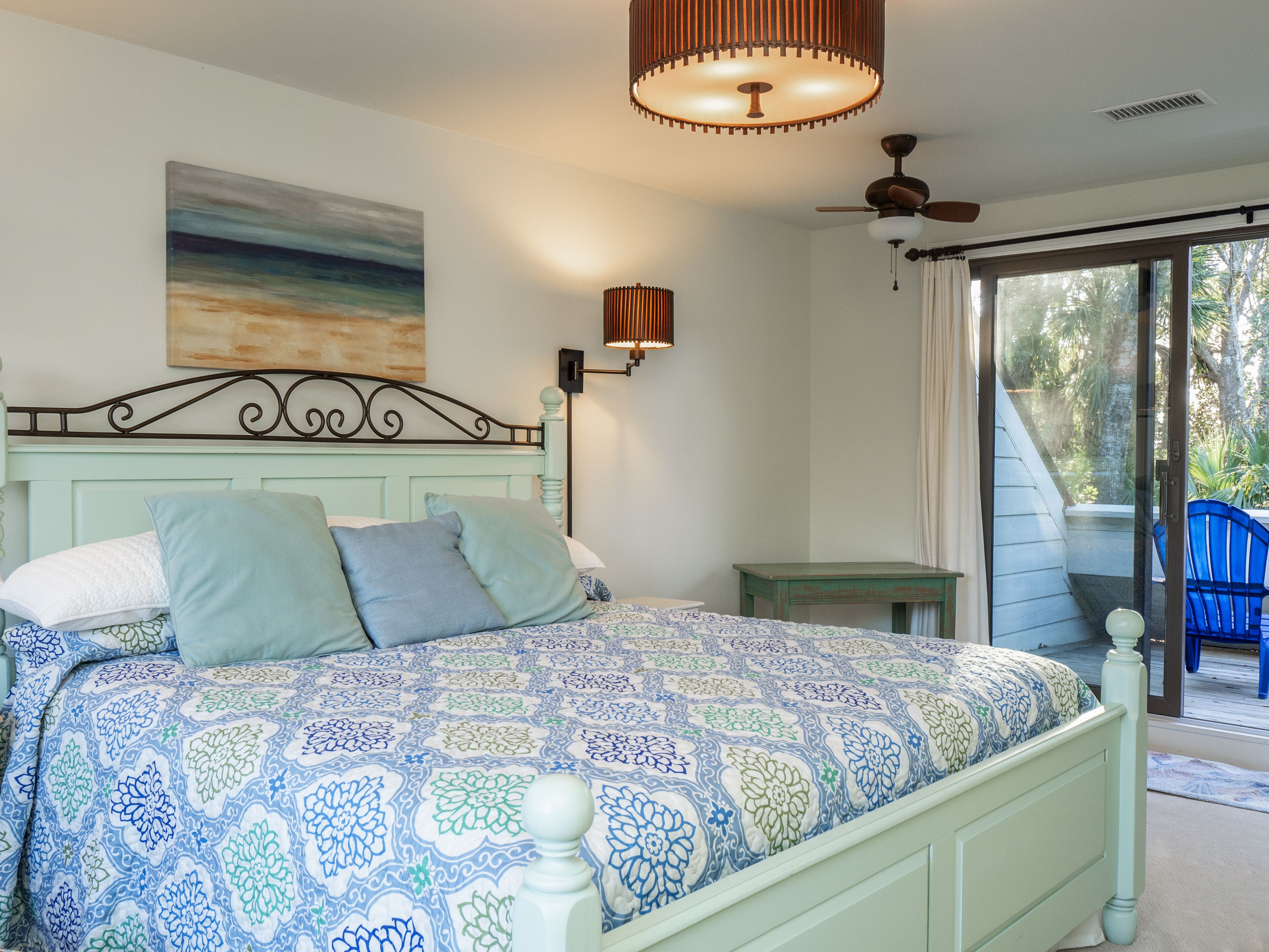 Quarto com cama, mesa de cabeceira, ventoinha no teto, janela com cortinas e vista para o exterior.