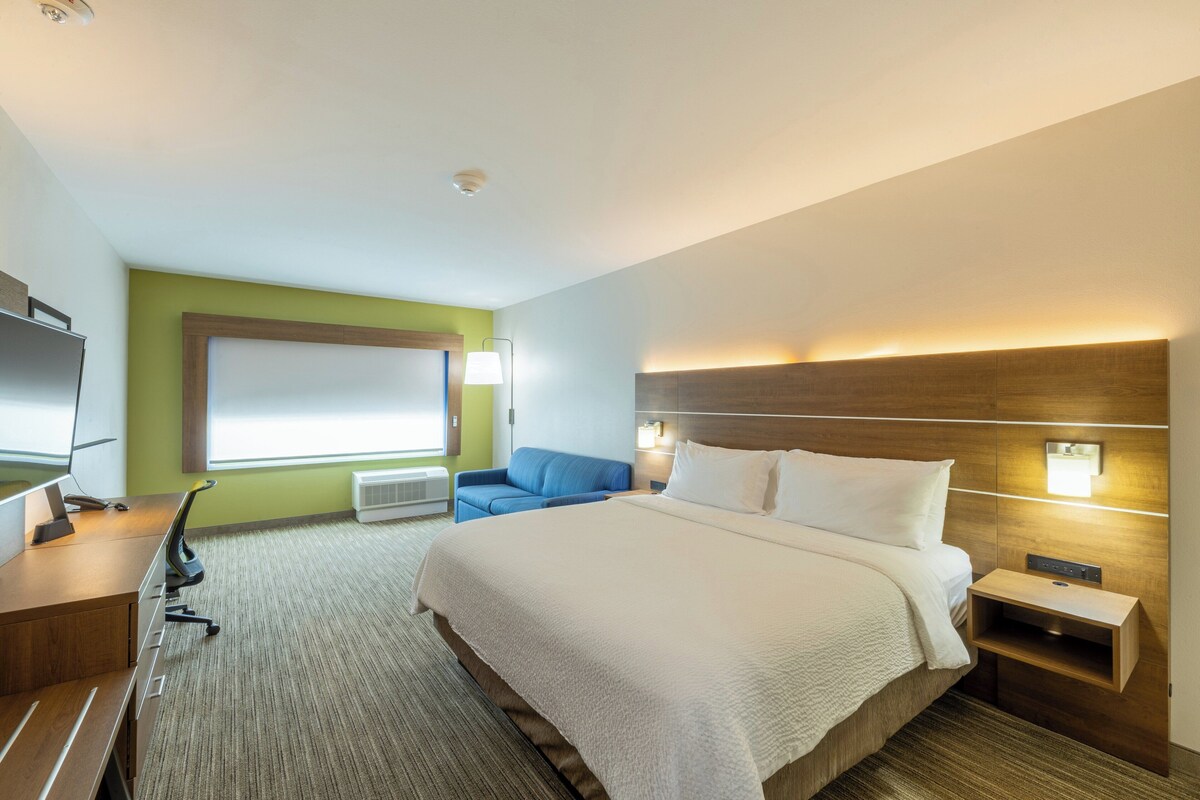A hotel room with a large bed, a desk with a computer, a blue chair, and a TV.