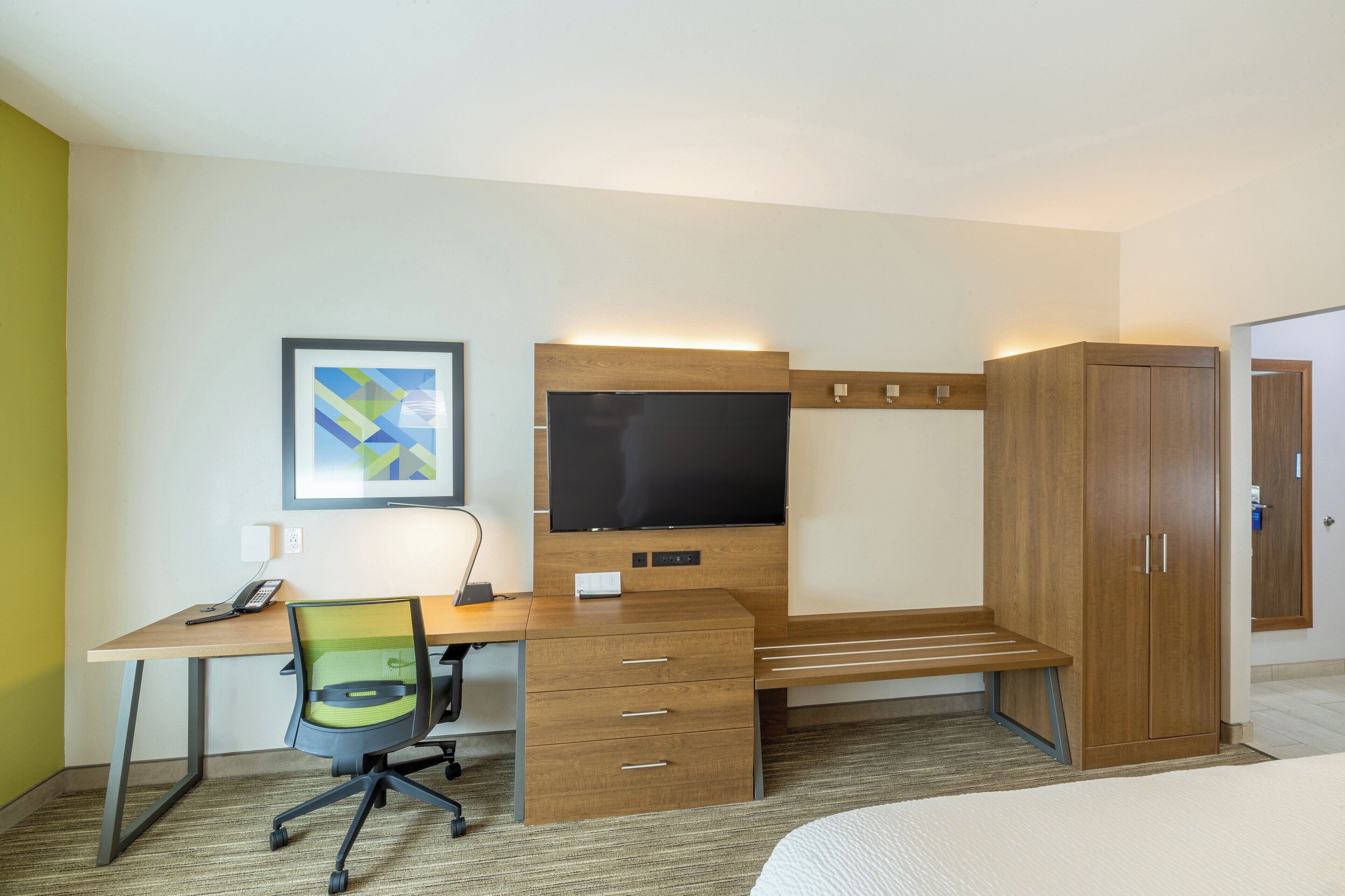 A hotel room with a wooden desk, a flat-screen TV, a chair, a bed, and a wardrobe.