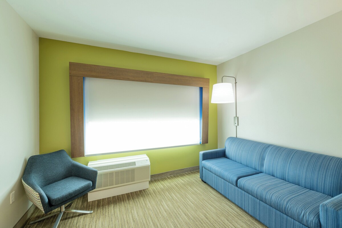 A room with a blue sofa, a blue armchair, a white air conditioner, and a large window with blinds.
