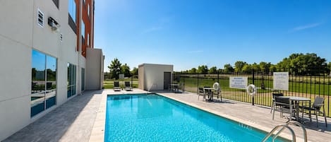A swimming pool with a clear blue water, surrounded by a deck area with chairs and tables, and a building with large windows and a sign indicating pool rules.
