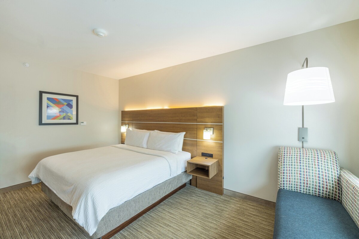 A hotel room with a bed, bedside table, lamp, and a small seating area with a patterned cushion.