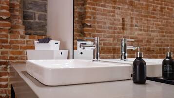 Un baño con un lavabo blanco, un espejo y una pared de ladrillo. Hay dos botellas de champú y una toalla en la encimera.