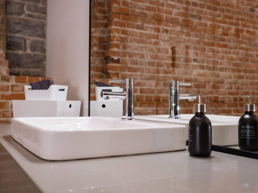 Un baño con un lavabo blanco, un espejo y una pared de ladrillo. Hay dos botellas de champú y una toalla en la encimera.