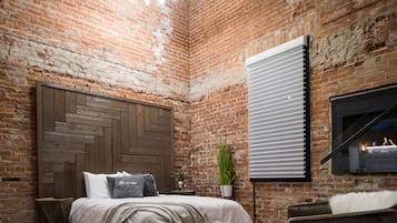 Un dormitorio con paredes de ladrillo visto, cabecero de madera, cama con ropa de cama blanca, mesita de noche con una planta y chimenea.