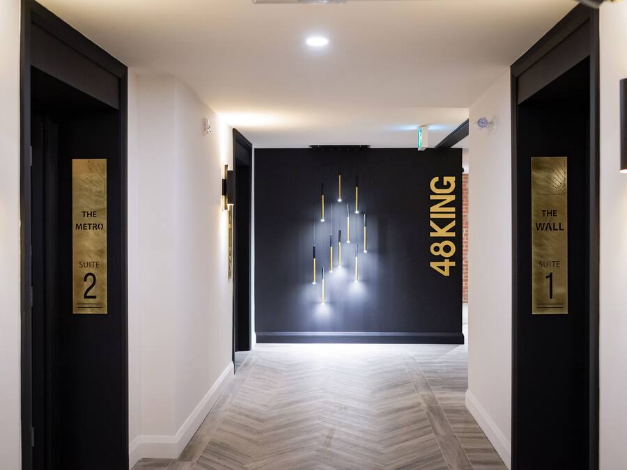 Um corredor moderno de hotel com paredes em preto e branco, sinalização dourada para 'The Metro Suite 2' e 'The Wall Suite 1', e piso de madeira clara.