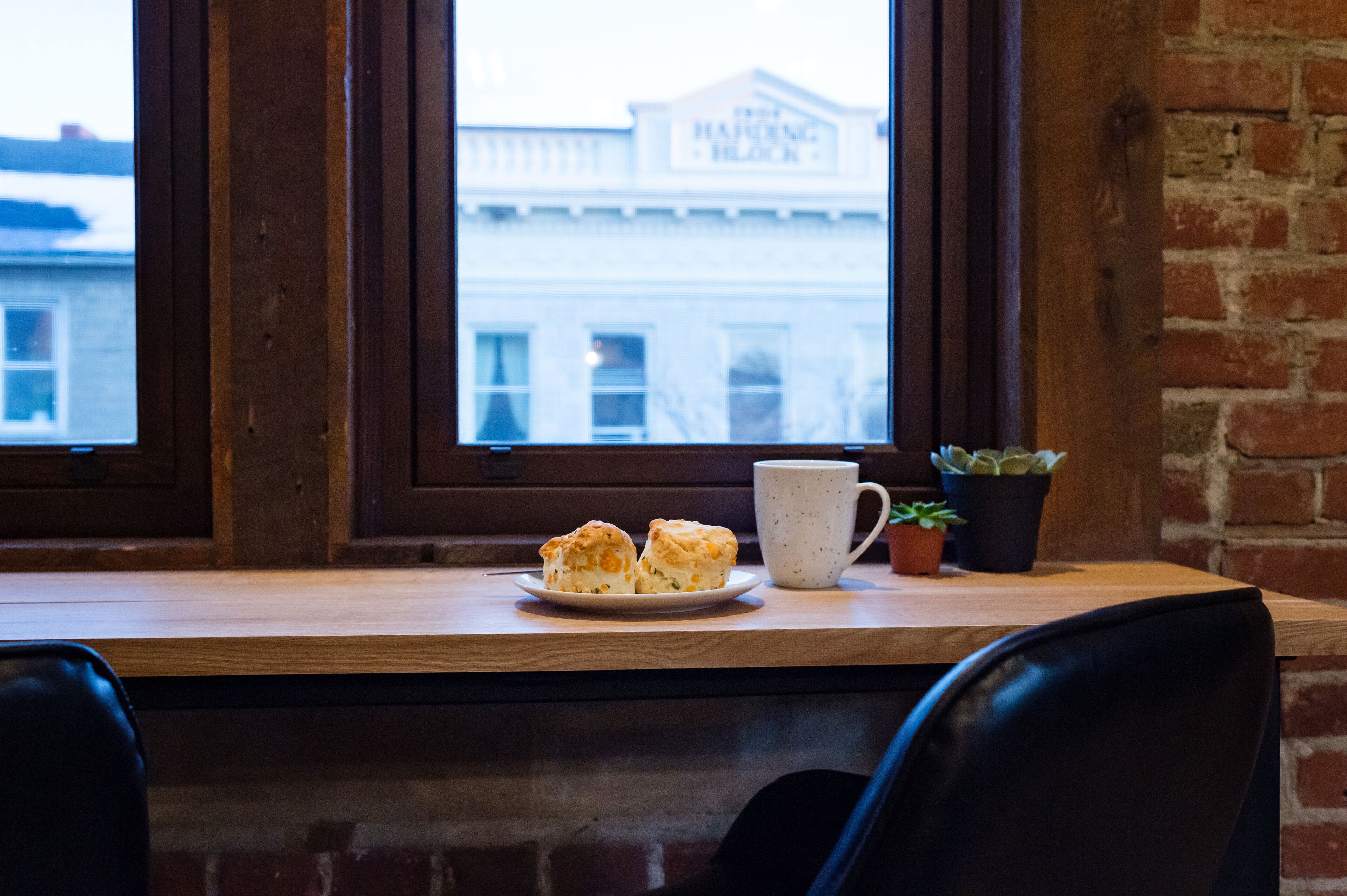 A table with a plate of pastries and a cup, a small potted plant, and a view of a building through a window.
