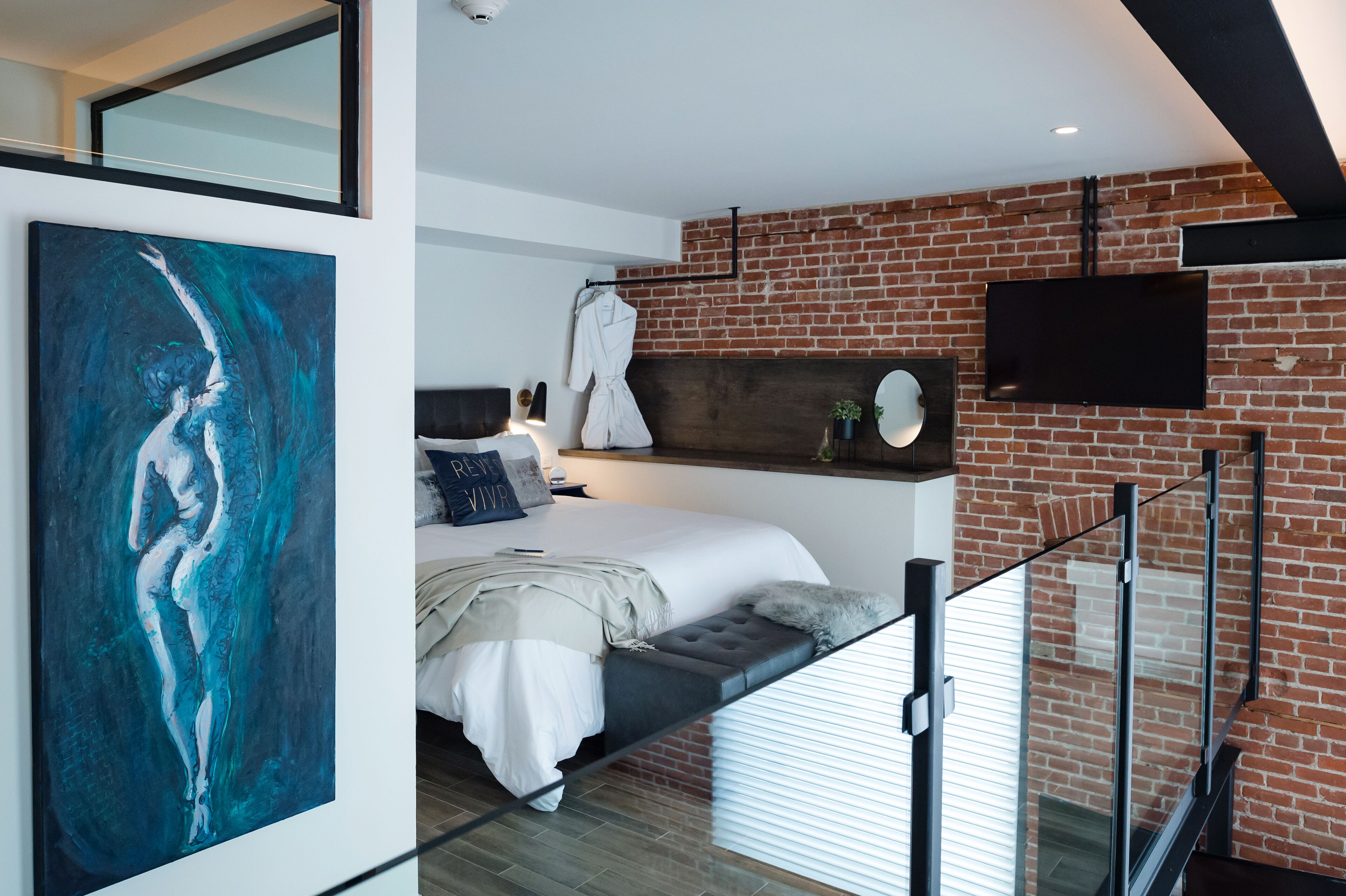 Un dormitorio moderno con pared de ladrillo, un cuadro grande, una cama con cabecero y un televisor montado.