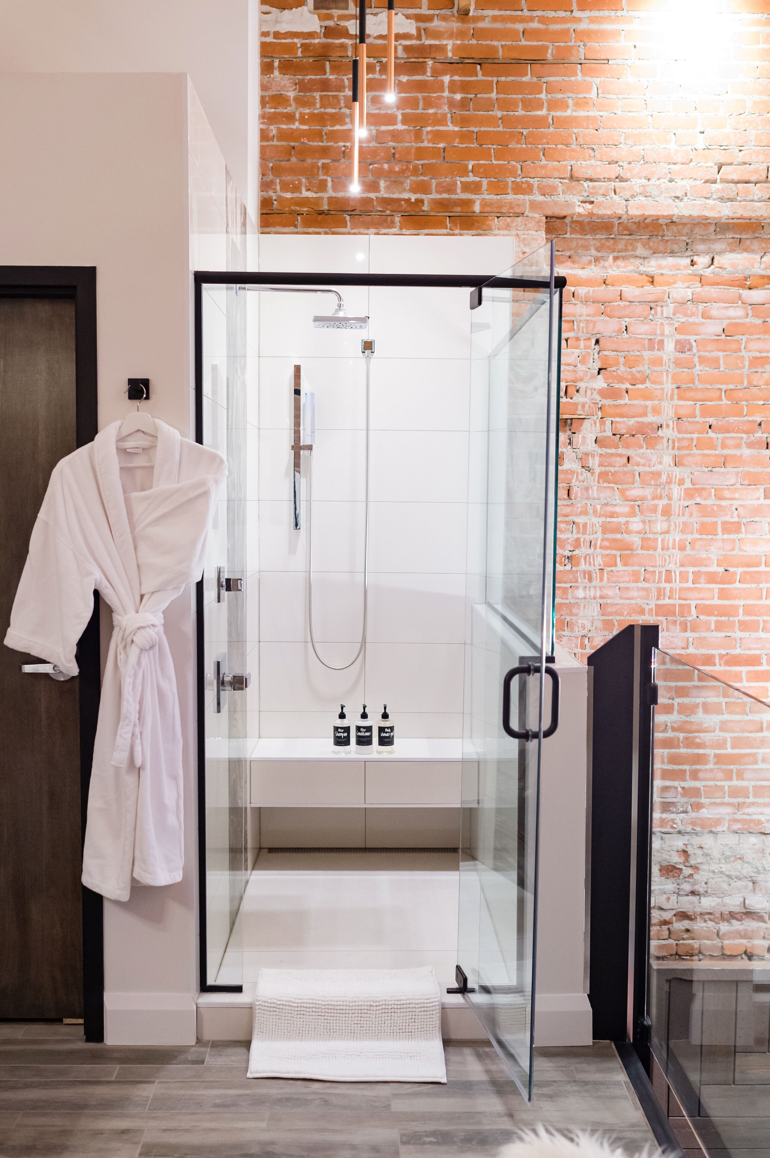 A modern bathroom with a glass-enclosed shower, a white robe hanging on the door, and a brick wall.
