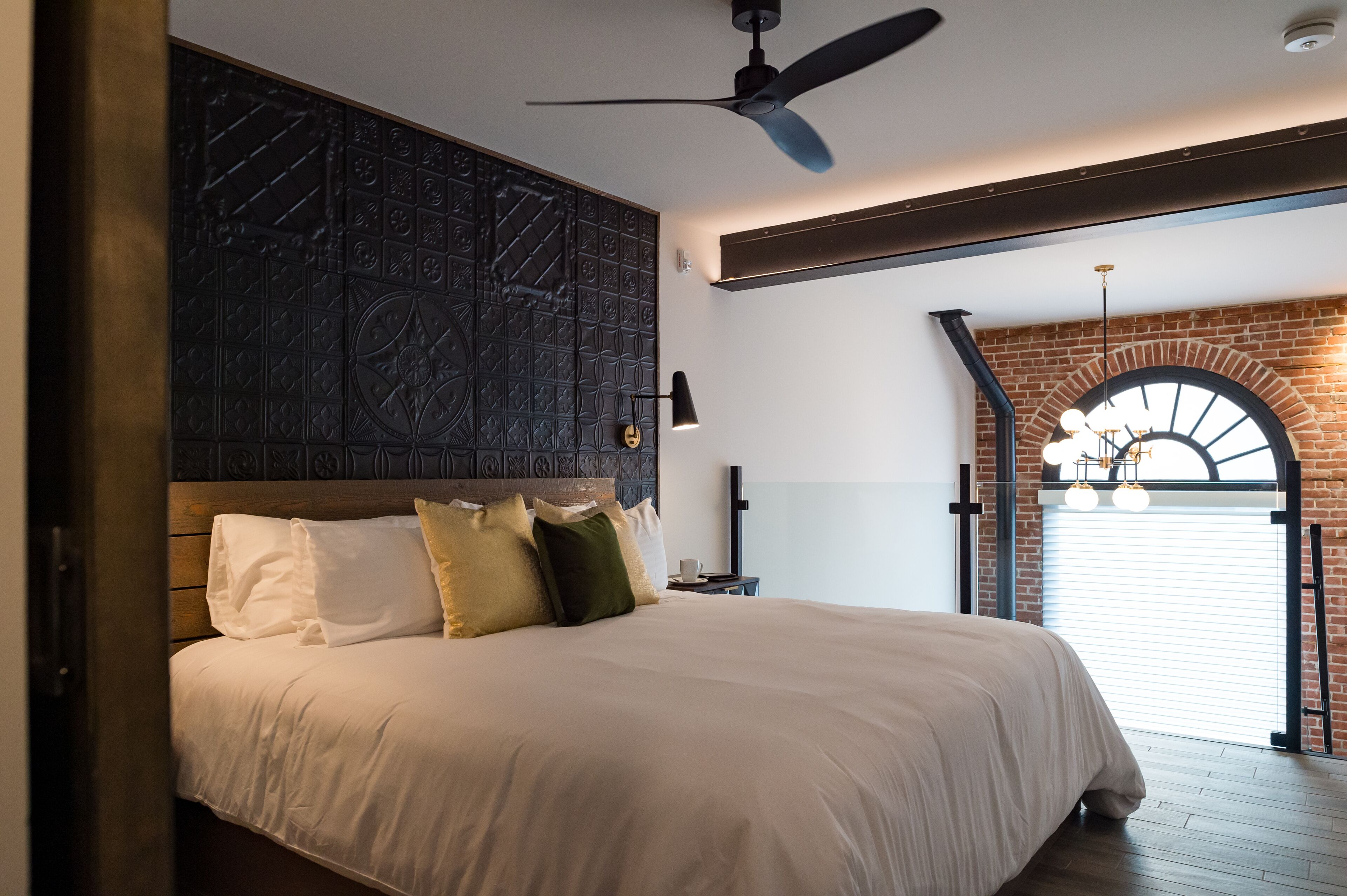 A bedroom with a large bed, a ceiling fan, a brick wall, and a window with a view.
