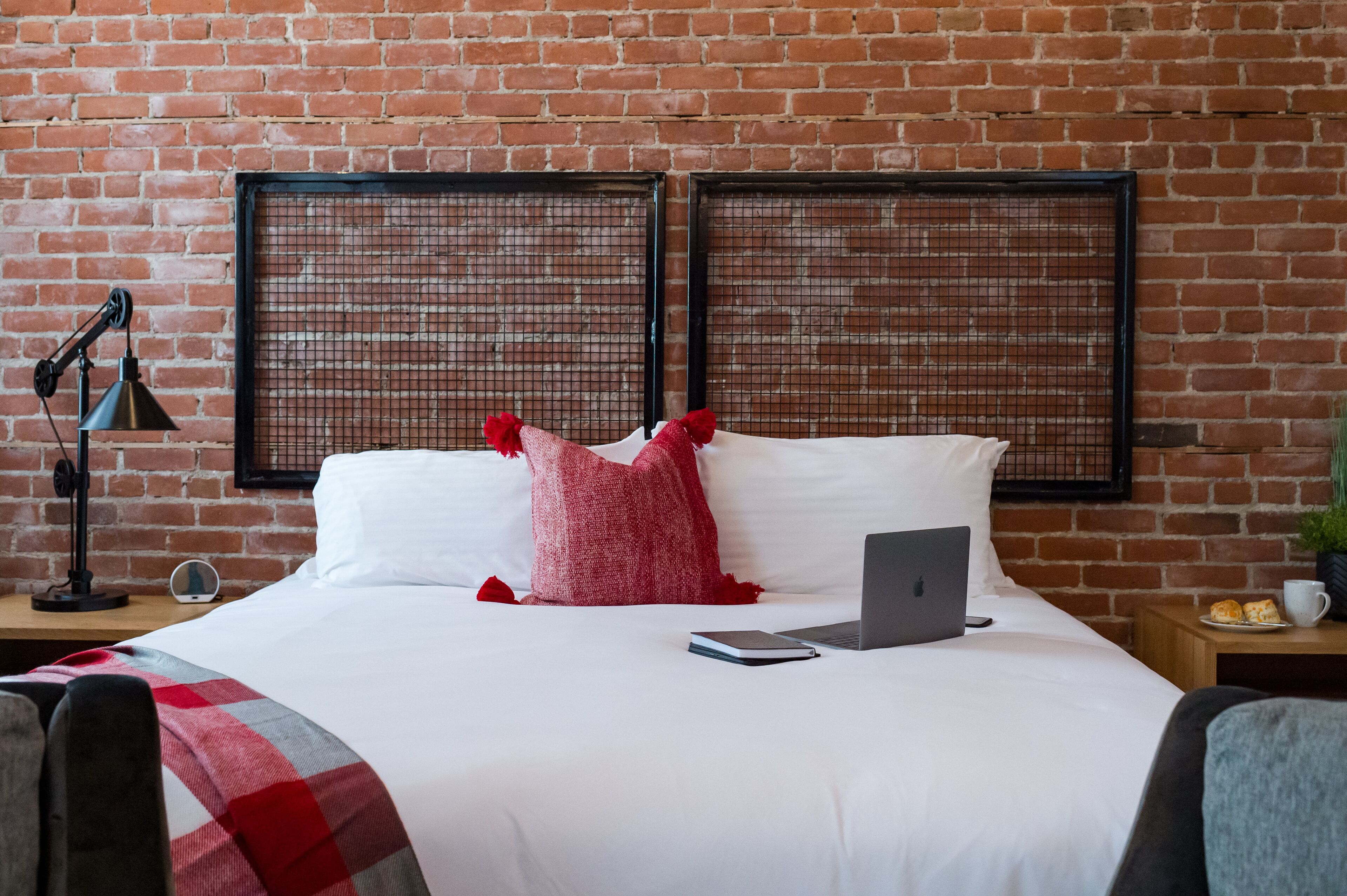 A hotel room with a brick wall, a bed with white bedding, a laptop, a bedside lamp, and a small table with a cup and pastries.