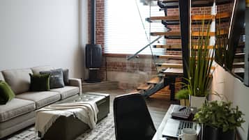 Una sala de estar moderna con un sofá, un reposapiés y una mesa de centro. Se ve un escritorio con una laptop, una taza y una planta. En el fondo, se observa una escalera con barandilla de madera.