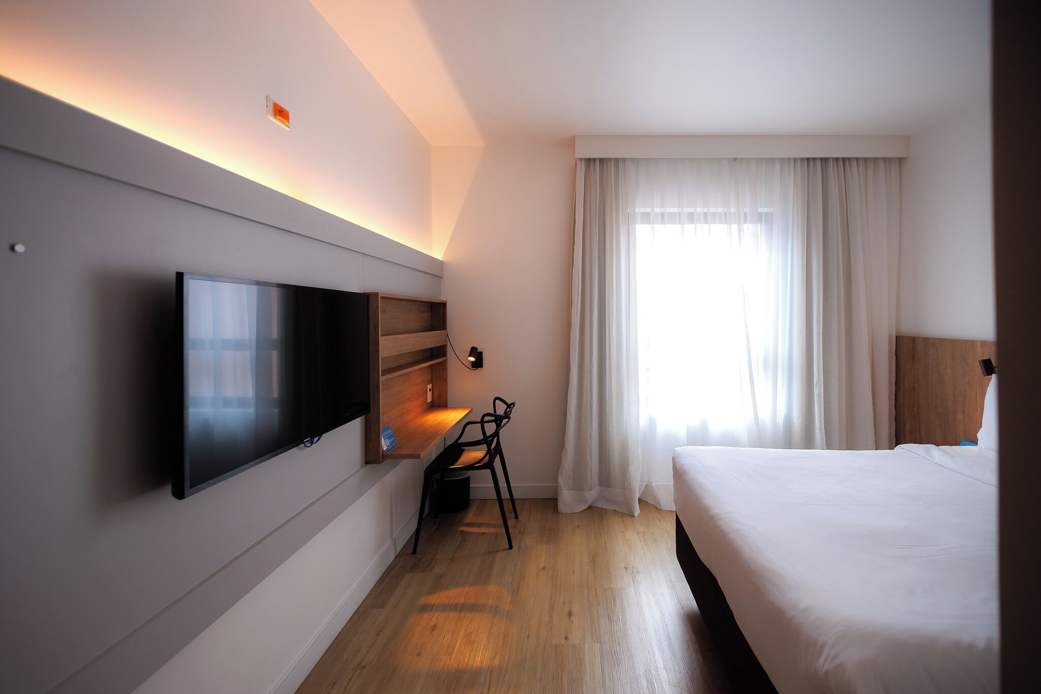 Een hotelkamer met een flatscreen televisie, een bureau met stoel, een bed en een groot raam met gordijnen.