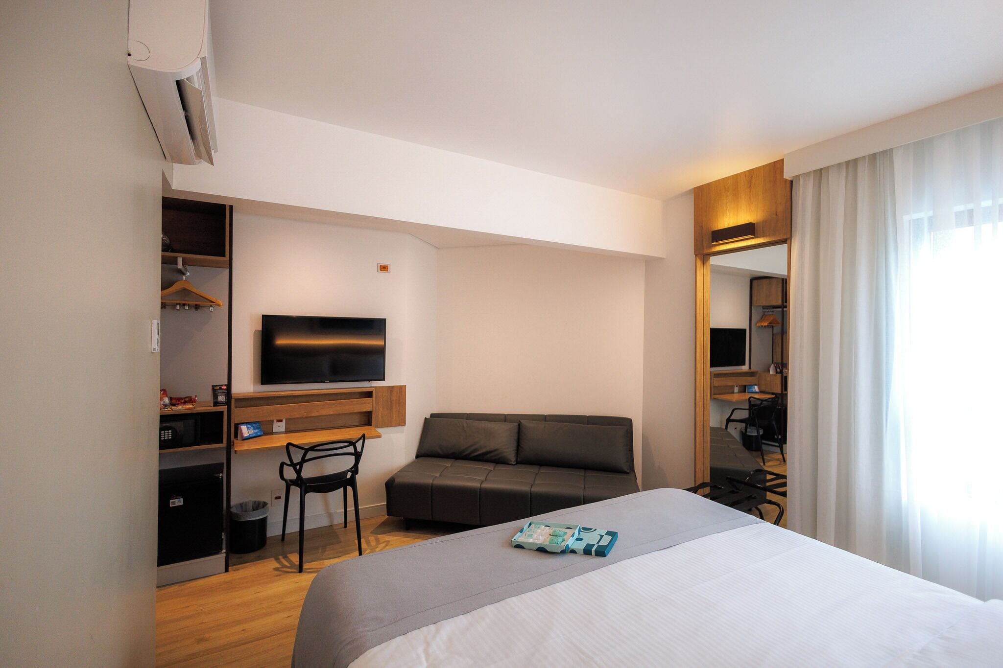 A modern hotel room with a bed, a sofa, a desk with a flat-screen TV, a chair, and a wardrobe.