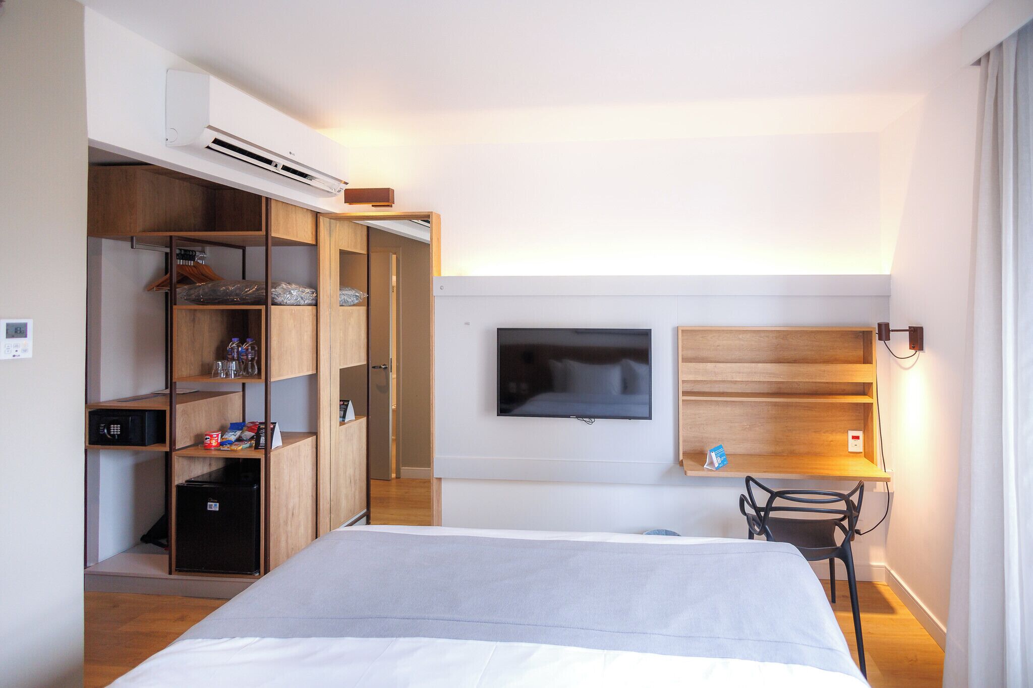 Um quarto com cama, televisão na parede, escrivaninha com cadeira e guarda-roupa com prateleiras.