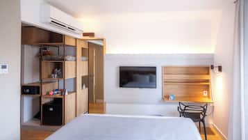 Um quarto com cama, televisão na parede, escrivaninha com cadeira e guarda-roupa com prateleiras.