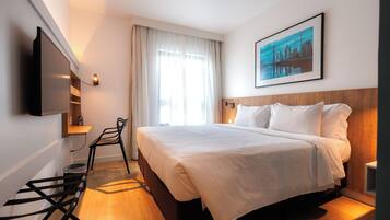 Quarto de hotel com cama, televisão fixada na parede, janela com cortinas e um quadro na parede.