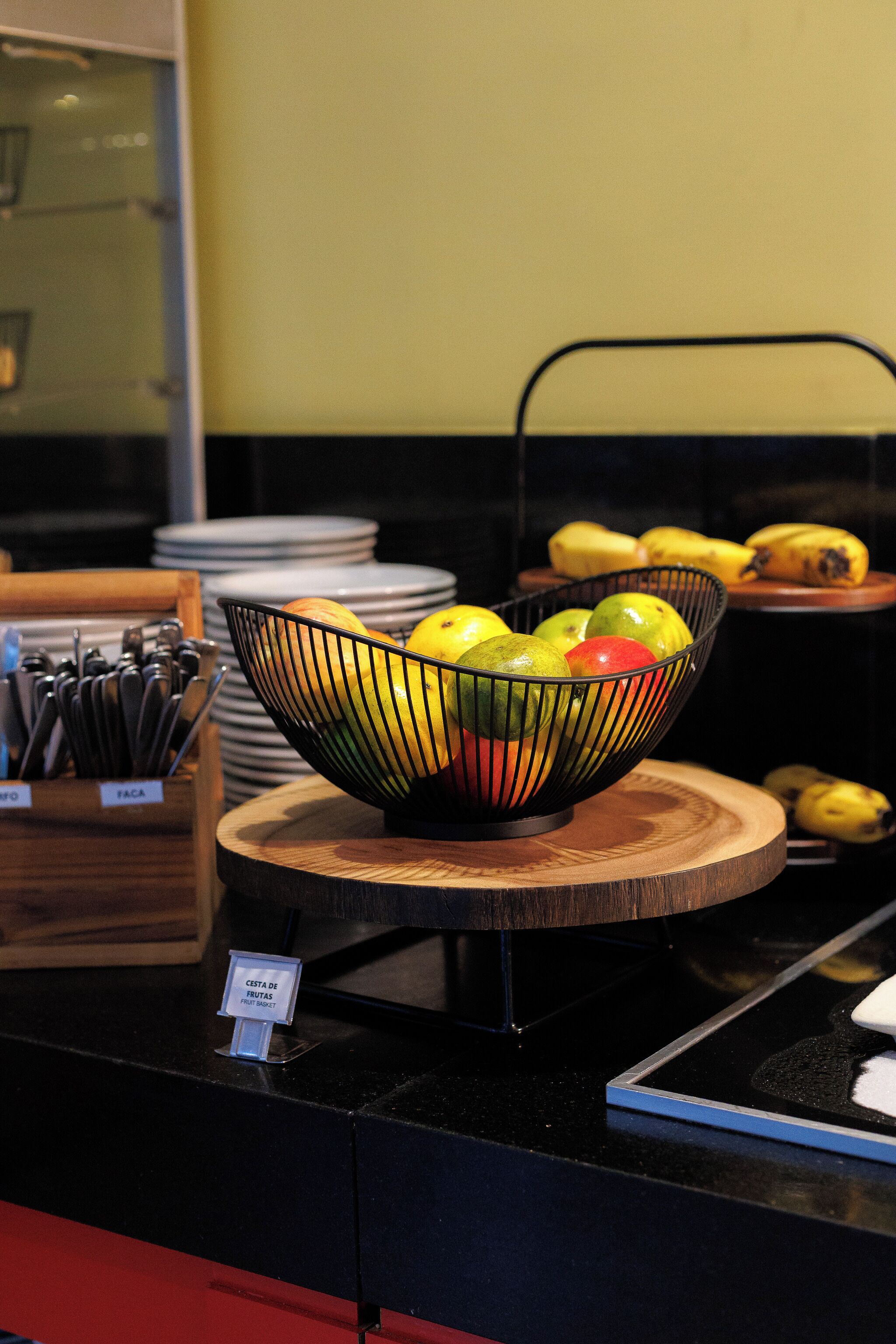 Un frutero sobre una tabla de madera, una pila de platos y un letrero que indica un desayuno tipo bufé.