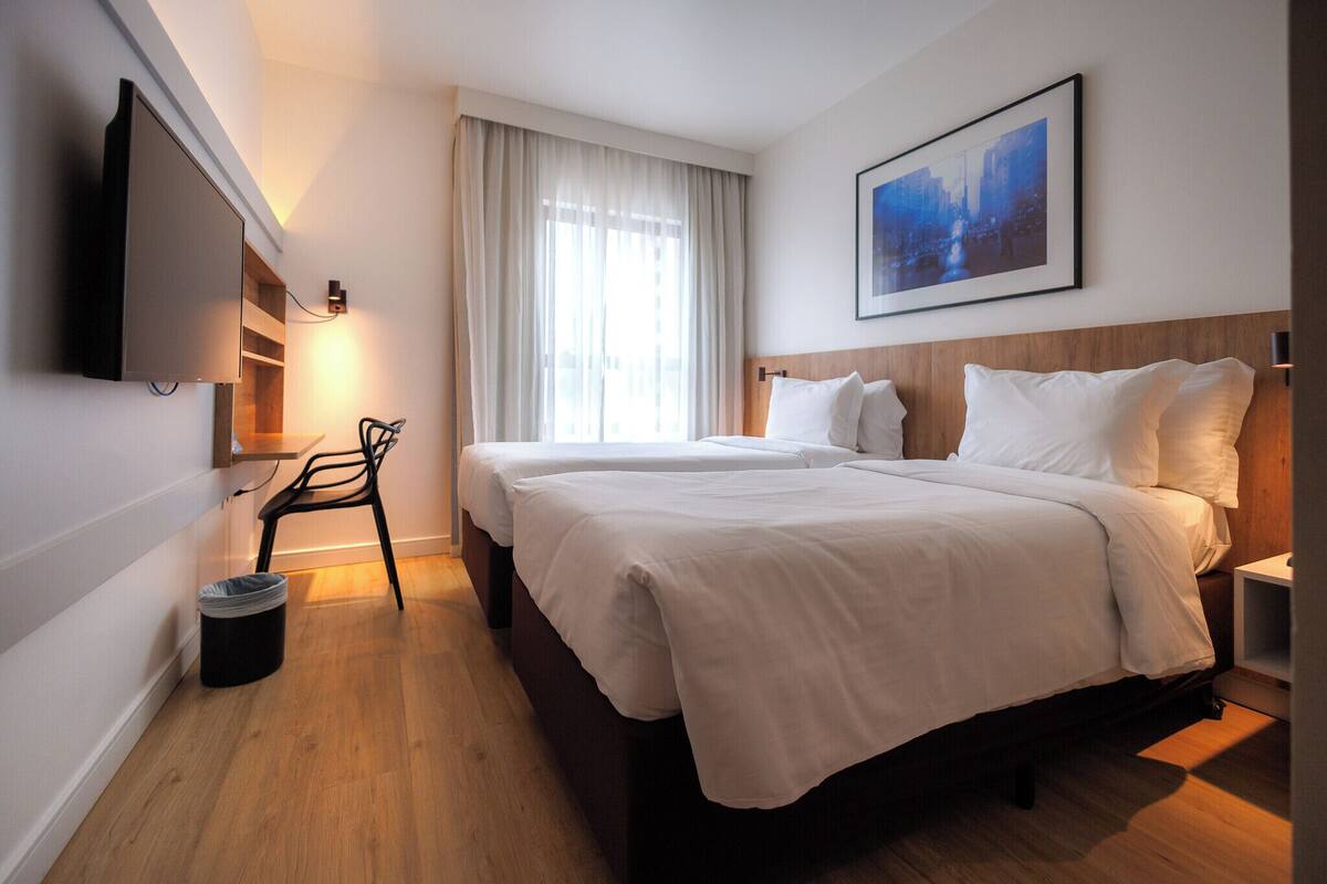 Une chambre d’hôtel avec un lit, un téléviseur fixé au mur, une chaise, une poubelle et un tableau accroché au mur.
