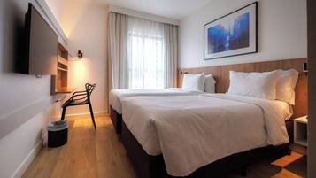 Quarto de hotel com cama, televisão fixada na parede, uma cadeira, um cesto de lixo e um quadro na parede.