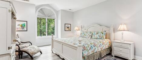 A bedroom with a large bed, two bedside tables, a ceiling fan, and a window with a view of trees.