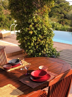 A wooden deck with a dining table set for two, surrounded by lush greenery and a pool.