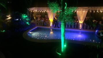 Zona de piscina al aire libre con escenario y asientos, rodeada de follaje e iluminada por luces colgantes.