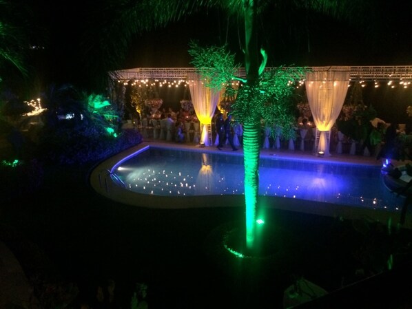 Zona de piscina al aire libre con escenario y asientos, rodeada de follaje e iluminada por luces colgantes.