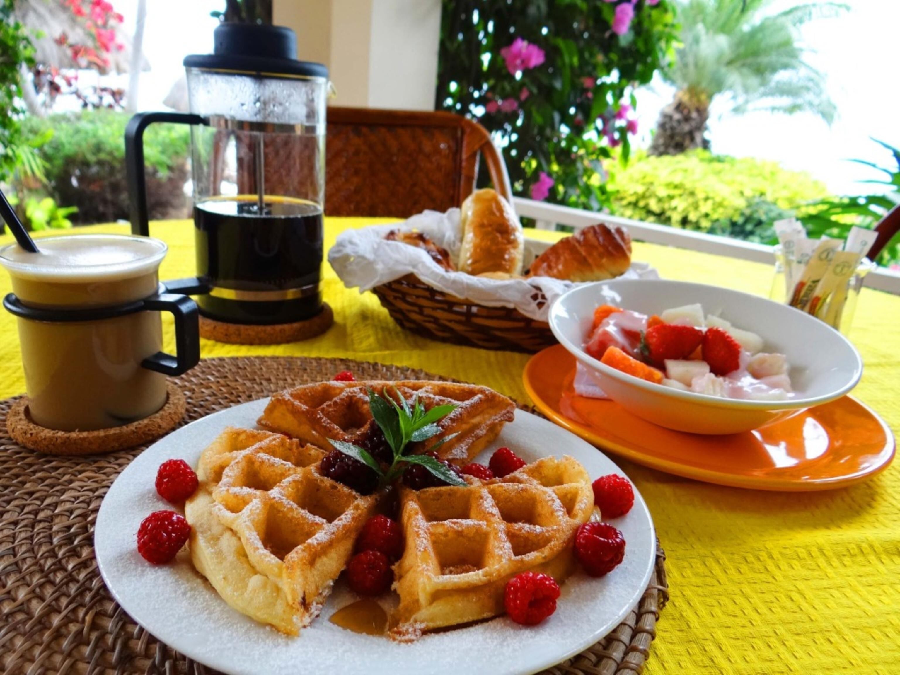 Un desayuno con waffles, una taza de café, un cuenco de fruta y una cesta de cruasanes sobre un mantel amarillo.