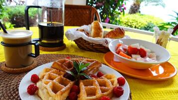 Un desayuno con waffles, una taza de café, un cuenco de fruta y una cesta de cruasanes sobre un mantel amarillo.