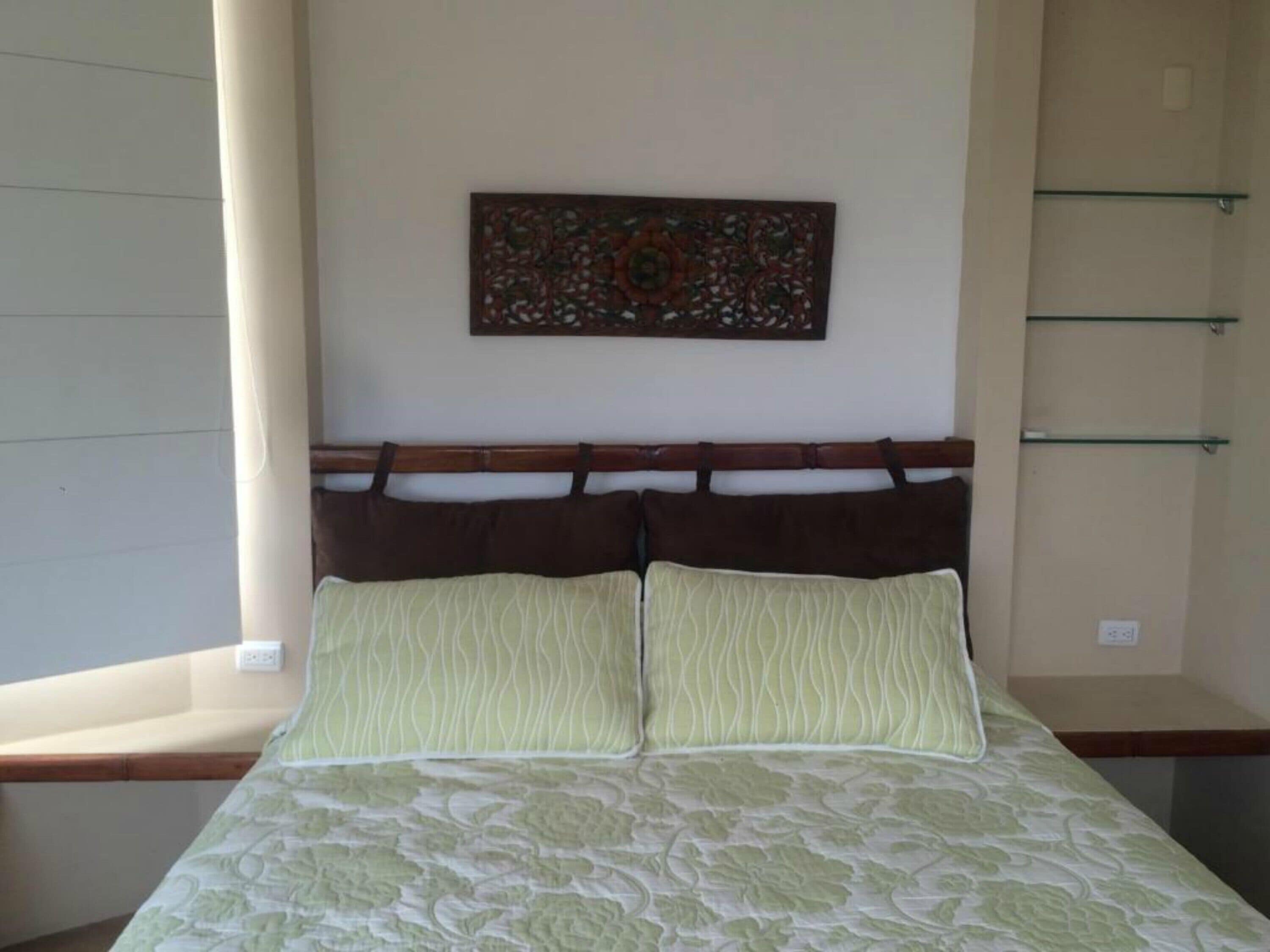 Un dormitorio con cama, cabecera, una obra de arte en la pared y una ventana con persianas.