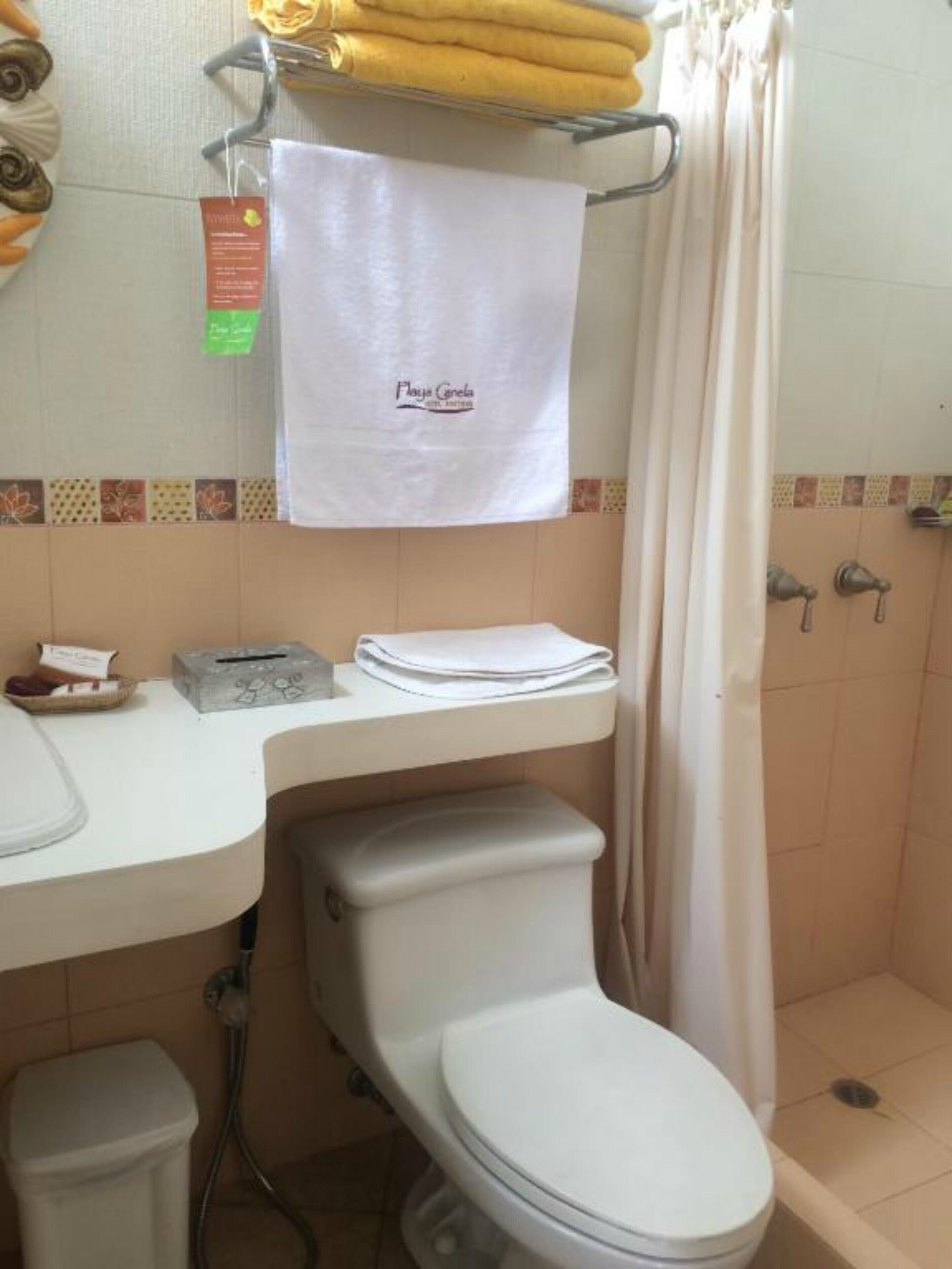 Un baño con inodoro, lavamanos y zona de ducha. Hay una toalla colgada en la pared y un cubo de basura pequeño en la encimera.