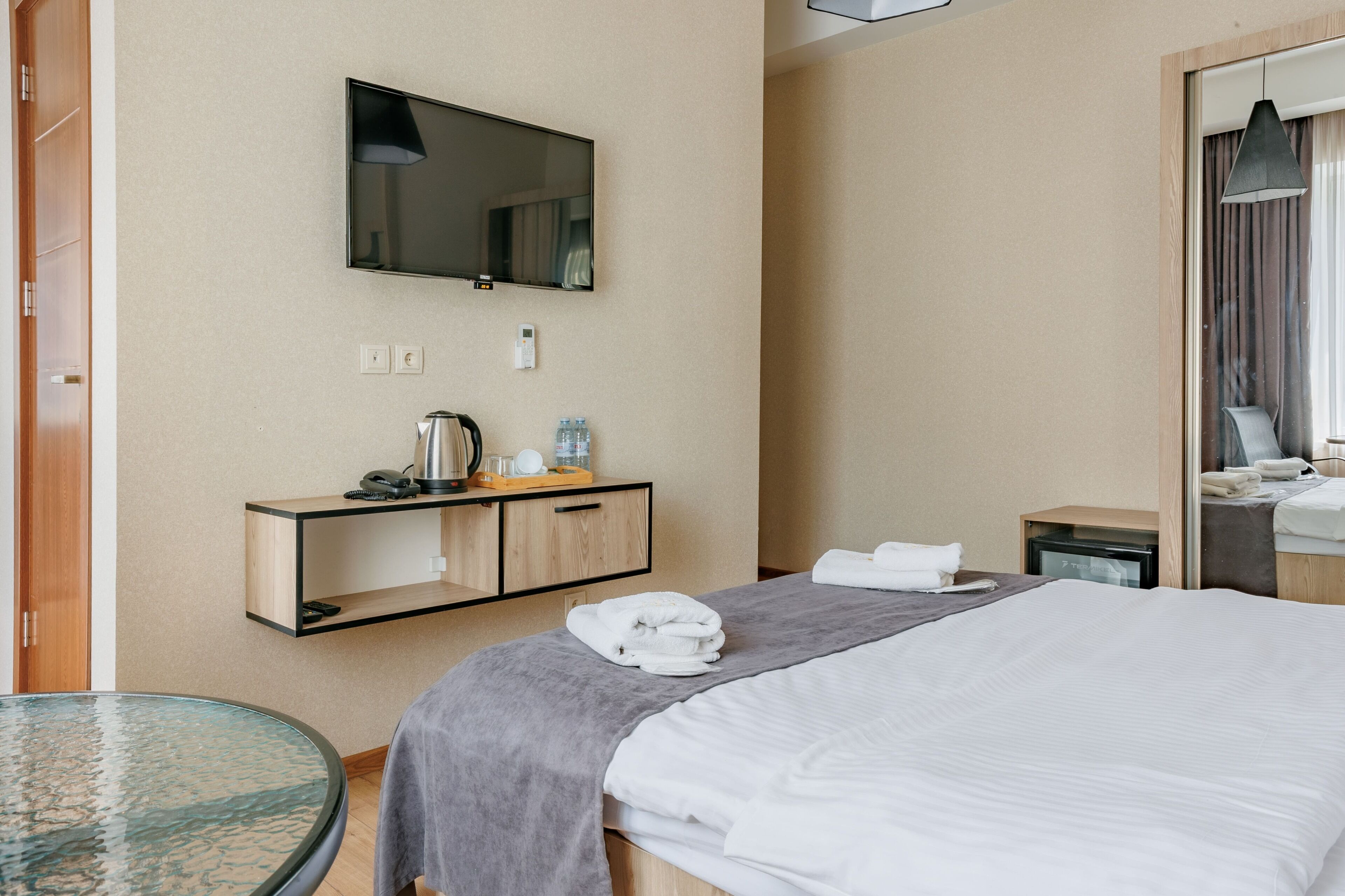A hotel room with a bed, a bedside table, a TV, a mirror, a small desk with a lamp, and a glass table.